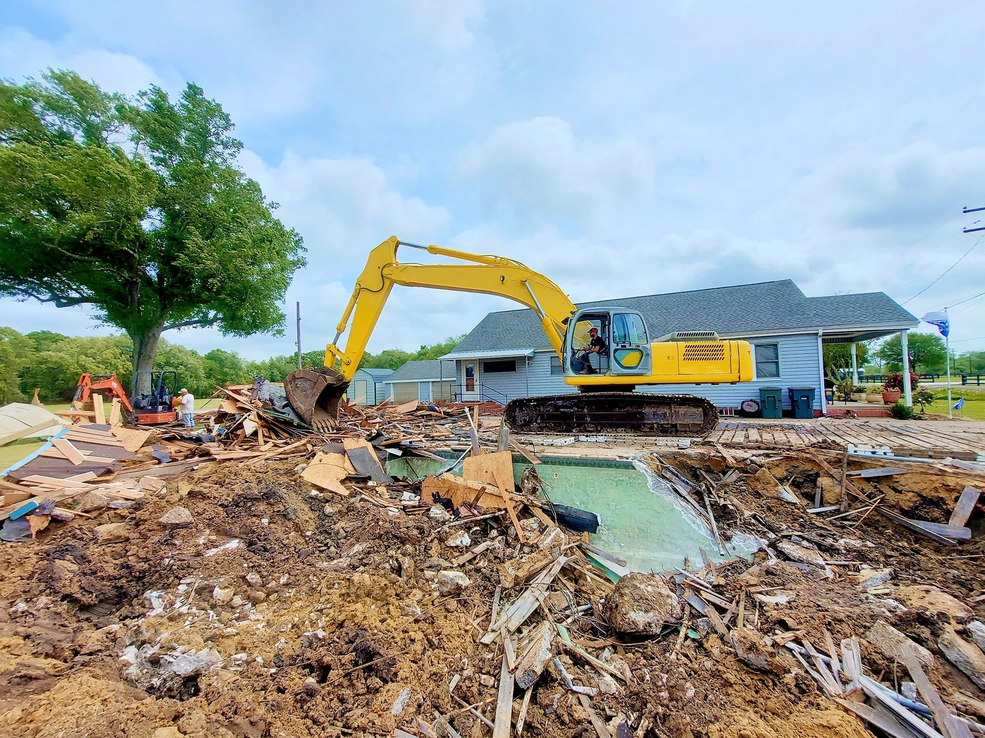 Large yellow excavator beside a demolished swimming pool and debris